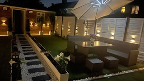 A well-lit outdoor patio area with a dining table and chairs, surrounded by a garden path and plants. - Heathrow Garden Lodge [Free Parking • Netflix • Prime • Coffee • WiFi] (Hayes)
