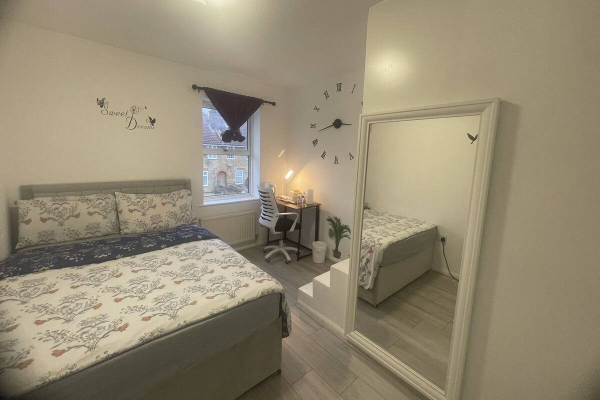 Een slaapkamer met een bed, een bureau, een stoel, een spiegel en een raam met uitzicht op een gebouw.