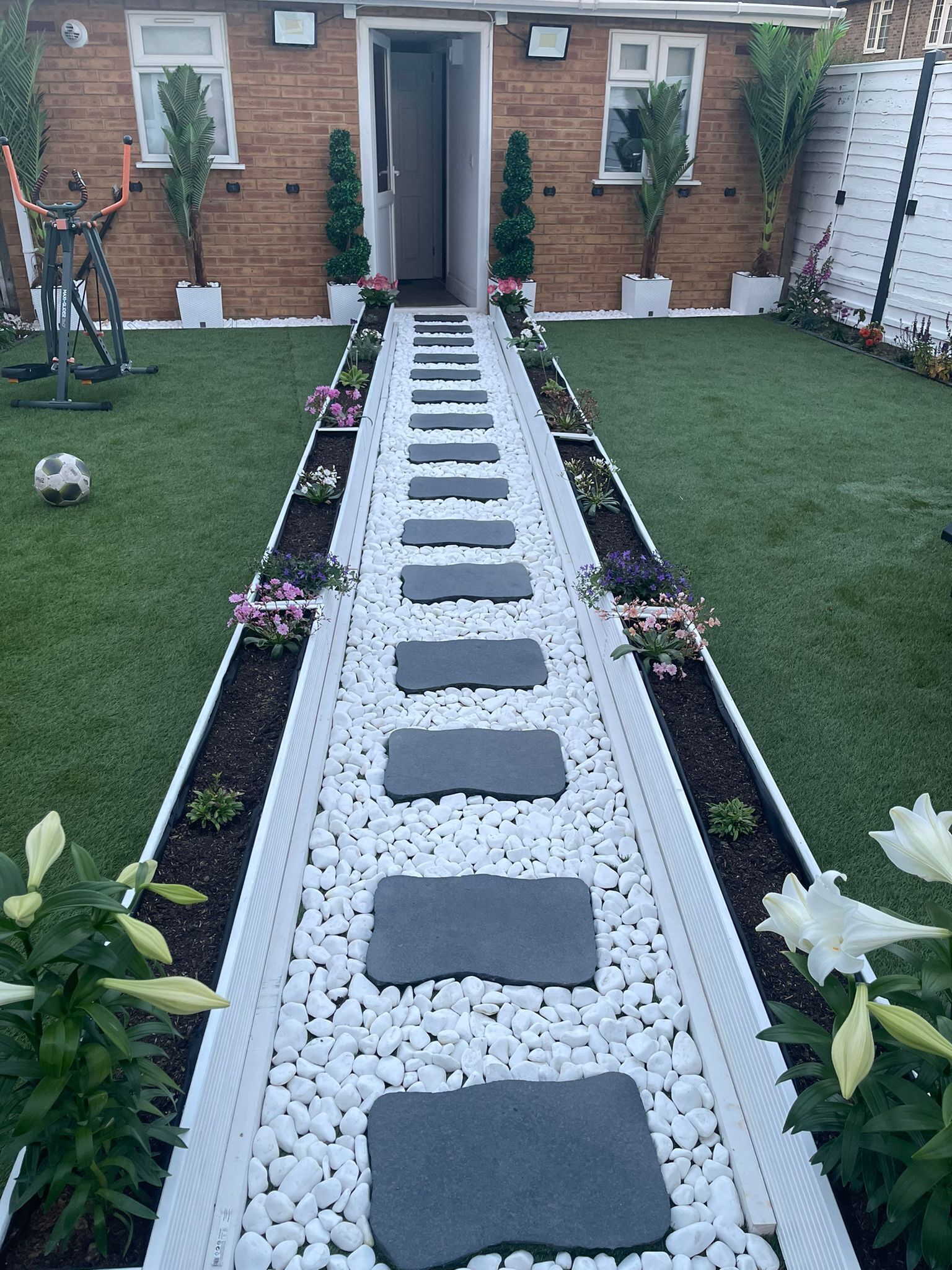 A garden pathway with stepping stones, bordered by white pebbles and plants, leading to a house with a brick facade and windows.