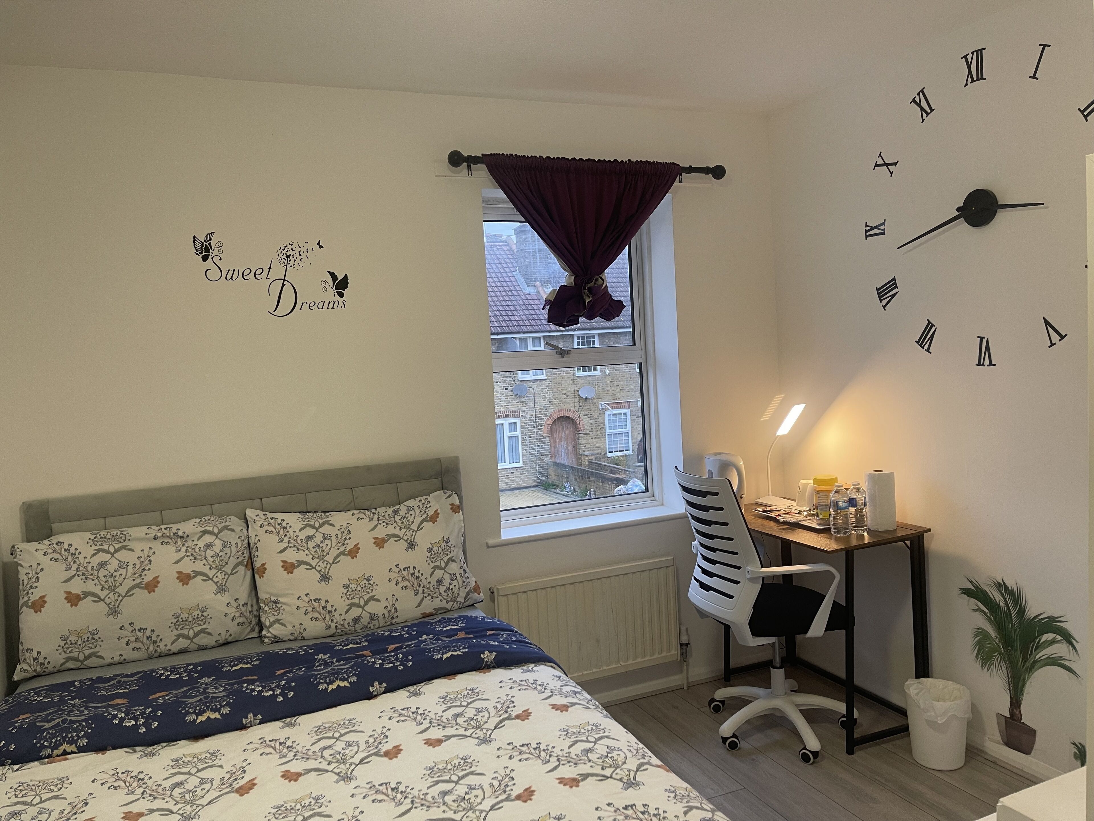 A bedroom with a bed, a desk, a chair, a window with curtains, a wall clock, and a wall decoration.