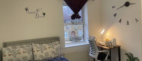 A bedroom with a bed, a desk, a chair, a window with curtains, a wall clock, and a wall decoration.