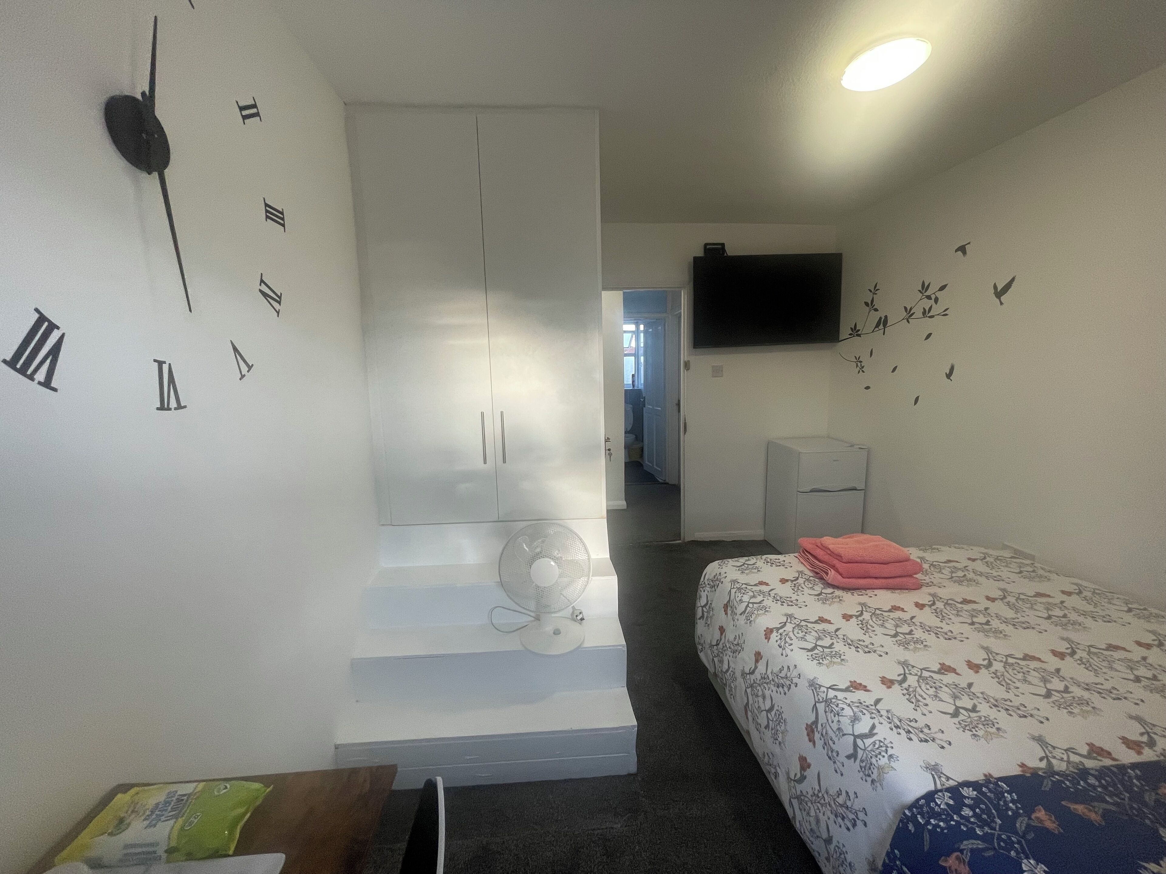 Ein Schlafzimmer mit einem Bett, einer Uhr, einem Ventilator und einem Fernseher an der Wand.