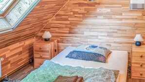Ein Schlafzimmer mit schrägen Wänden, Holzwänden, einem Bett mit gemusterten Betttextilien, zwei Nachttischen mit Lampen und gerahmten Bildern an der Wand.