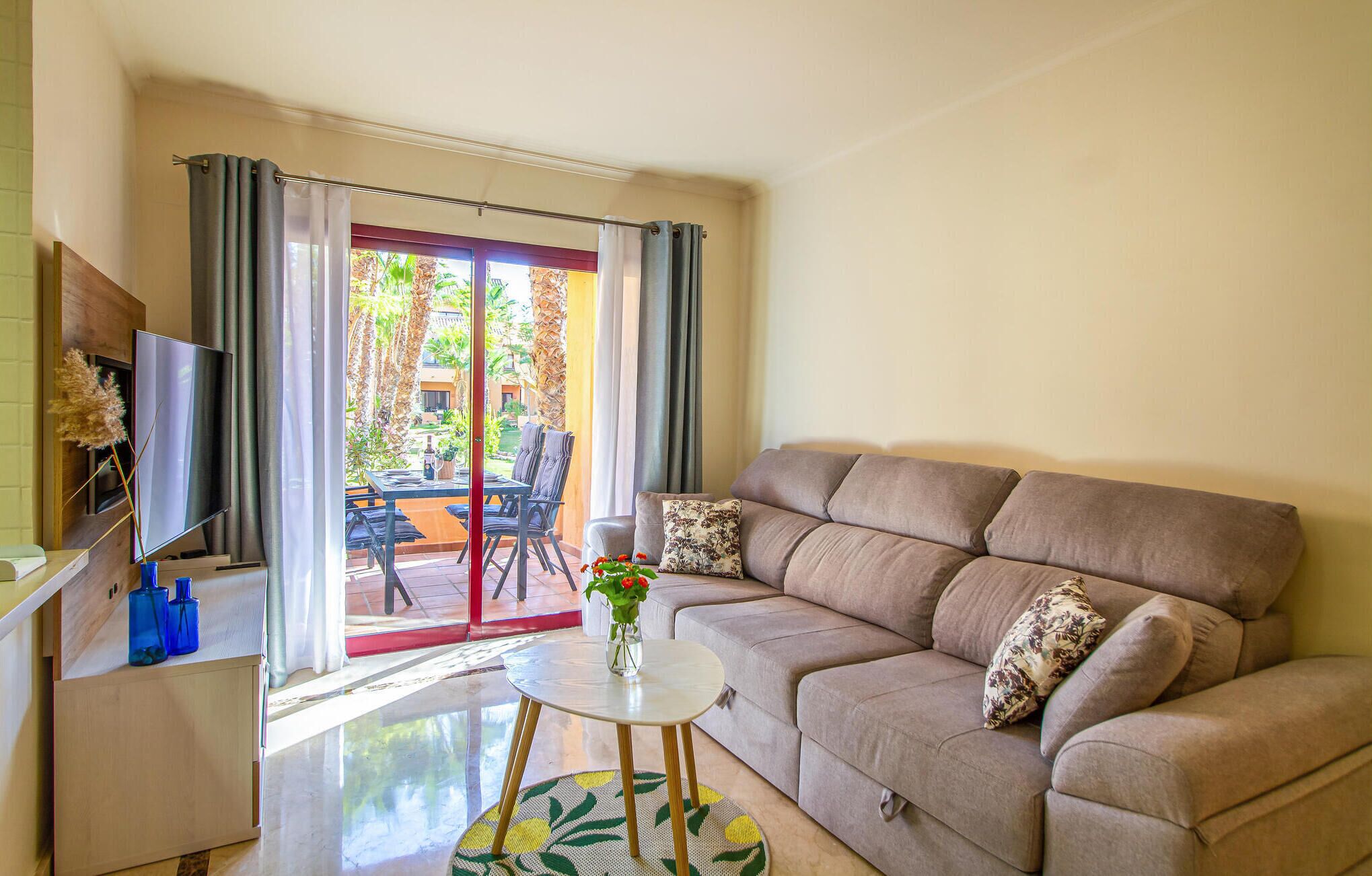 Una sala de estar con sofá, una mesita con un florero, un televisor sobre un mueble de madera y un ventanal corredero que da a un espacio exterior con mesa y sillas.
