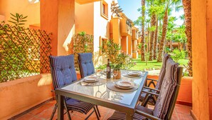 A dining area with a glass table, striped chairs, and a view of a courtyard with palm trees and a building in the background. - Beach front apartment in Los Alcázares (Los Alcázares)