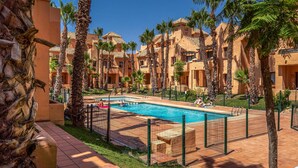 A resort with a swimming pool surrounded by palm trees and a building with a tiled roof. - Beach front apartment in Los Alcázares (Los Alcázares)