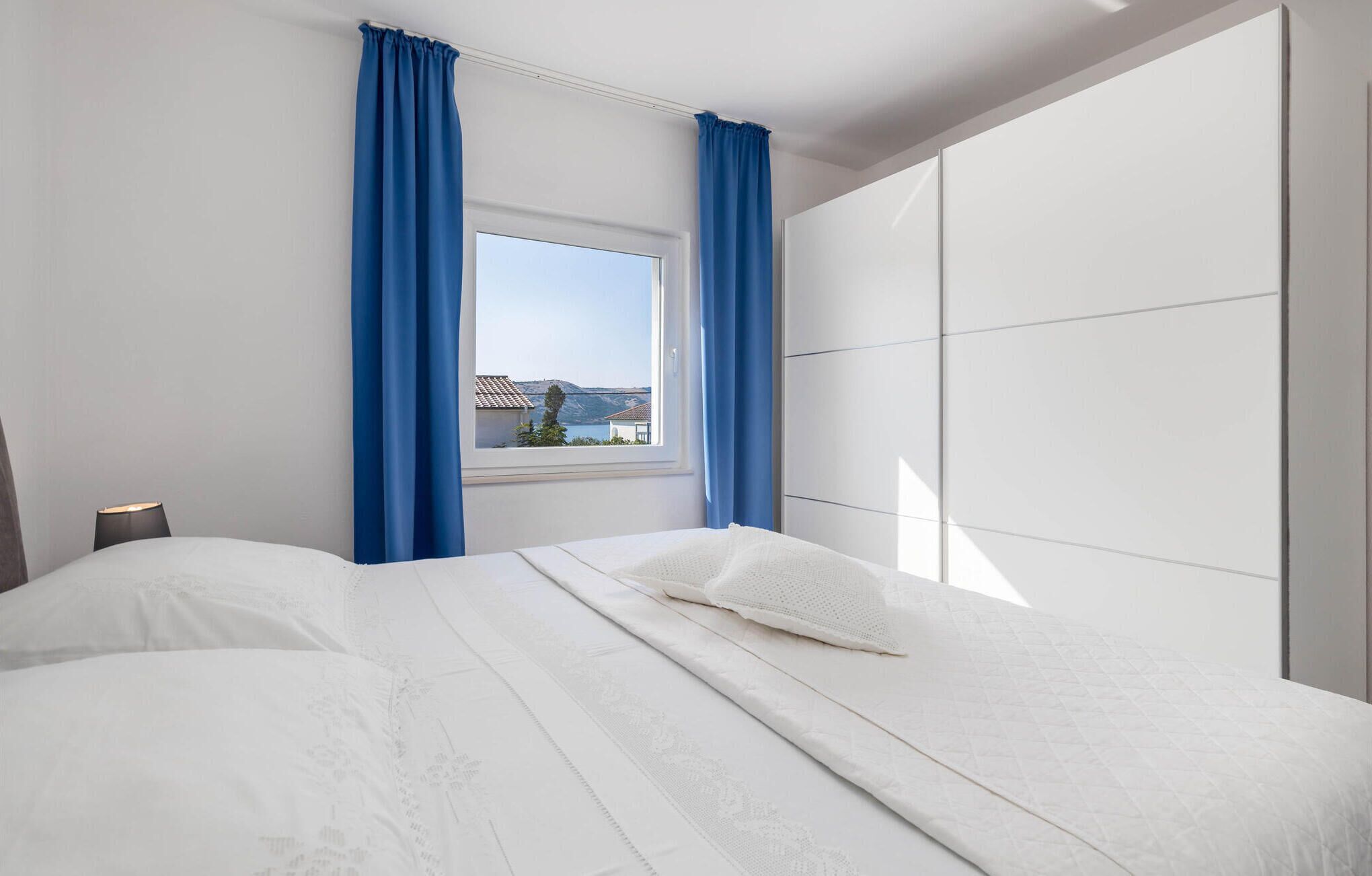 Une chambre avec un grand lit, une fenêtre avec des rideaux bleus et une vue sur la mer et les bâtiments.