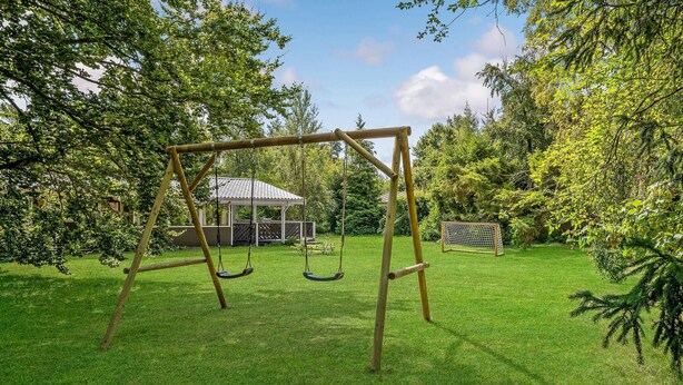 Ein hölzernes Schaukelgestell in einem grünen Garten mit einem Gazebo und einem Fußballtor im Hintergrund.