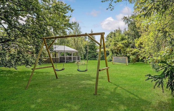 A wooden swing set in a grassy backyard with a gazebo and a soccer goal in the background.