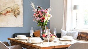 Ein Esstisch ist gedeckt mit weißen Tellern, klaren Weingläsern und einer Mittelstück-Anordnung aus Blumen und Früchten.