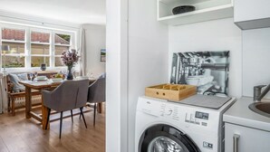 Eine moderne Küche mit integriertem Waschmaschine, ein Essbereich mit Holztisch und -stühlen sowie ein Ausblick ins Freie durch große Fenster.