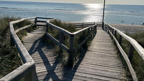 Ein hölzerner Steg führt zu einem Strand, im Hintergrund das Meer.