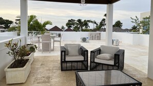 A covered outdoor seating area with wicker furniture and a glass table, overlooking a beach with palm trees and buildings in the background. - Rilaxing villa Ocean Village (Sosua)