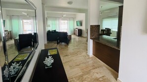 A spacious living area with a large mirror, a black coffee table with decorative items, and a kitchen area with a granite countertop and a window with blinds. - Rilaxing villa Ocean Village (Sosua)