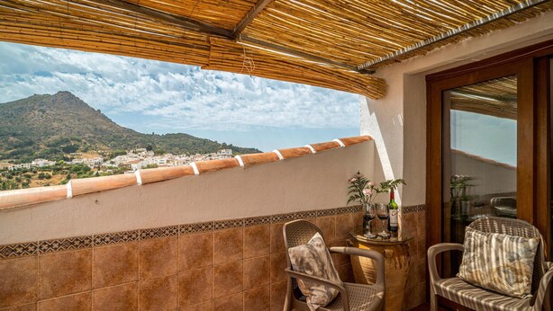 Ein Balkon mit Blick auf eine Stadt und ein Gebirge, Korbstühle, ein kleiner Tisch mit einer Vase und Weingläsern sowie eine Bambusdecke.