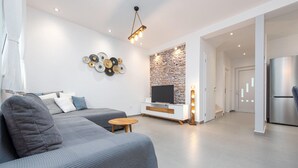 Ein modernes Wohnzimmer mit grauem Sofa, einem kleinen Holztisch und einem Fernseher an einem weißen Schrank vor einer Steinwand.