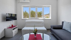 A modern room with a bed, a gray sofa, a TV, and a window with a view of trees and buildings. - Beautiful apartment in Murter with swimming pool (Murter)