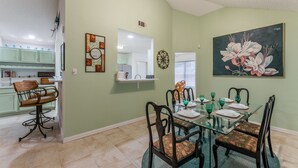 A dining area with a glass table and chairs, a painting on the wall, and a kitchen in the background. - Private Hot Tub, Stone Floors, Tall Fenced Yard, Fire Pit, & BBQ Grill (Grand Prairie)