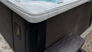 A hot tub with a wooden bench and a covered area, set outdoors with a brick wall and a wooden fence in the background. - Private Hot Tub, Stone Floors, Tall Fenced Yard, Fire Pit, & BBQ Grill (Grand Prairie)