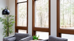 A modern living room with two grey armchairs, a small round table, and a floor lamp. Large windows offer a view of a forested area.
