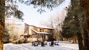A rustic wooden cabin with a deck and outdoor seating area, surrounded by trees and snow.