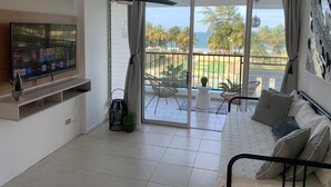 Een moderne woonkamer met een televisie, een plafondventilator, een balkon met uitzicht op zee en palmbomen, en een bank met kussens.
