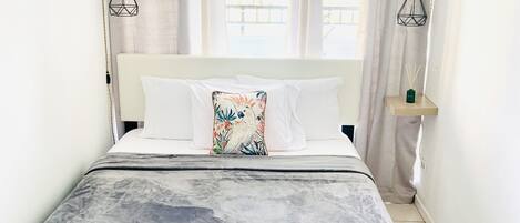 Una cama bien hecha con un edredón gris, almohadas blancas y una almohada floral colorida. Hay una mesita de noche con un jarrón verde y una lámpara. La habitación tiene paredes blancas, una ventana con cortinas y un equipo de aire acondicionado en la pared.
