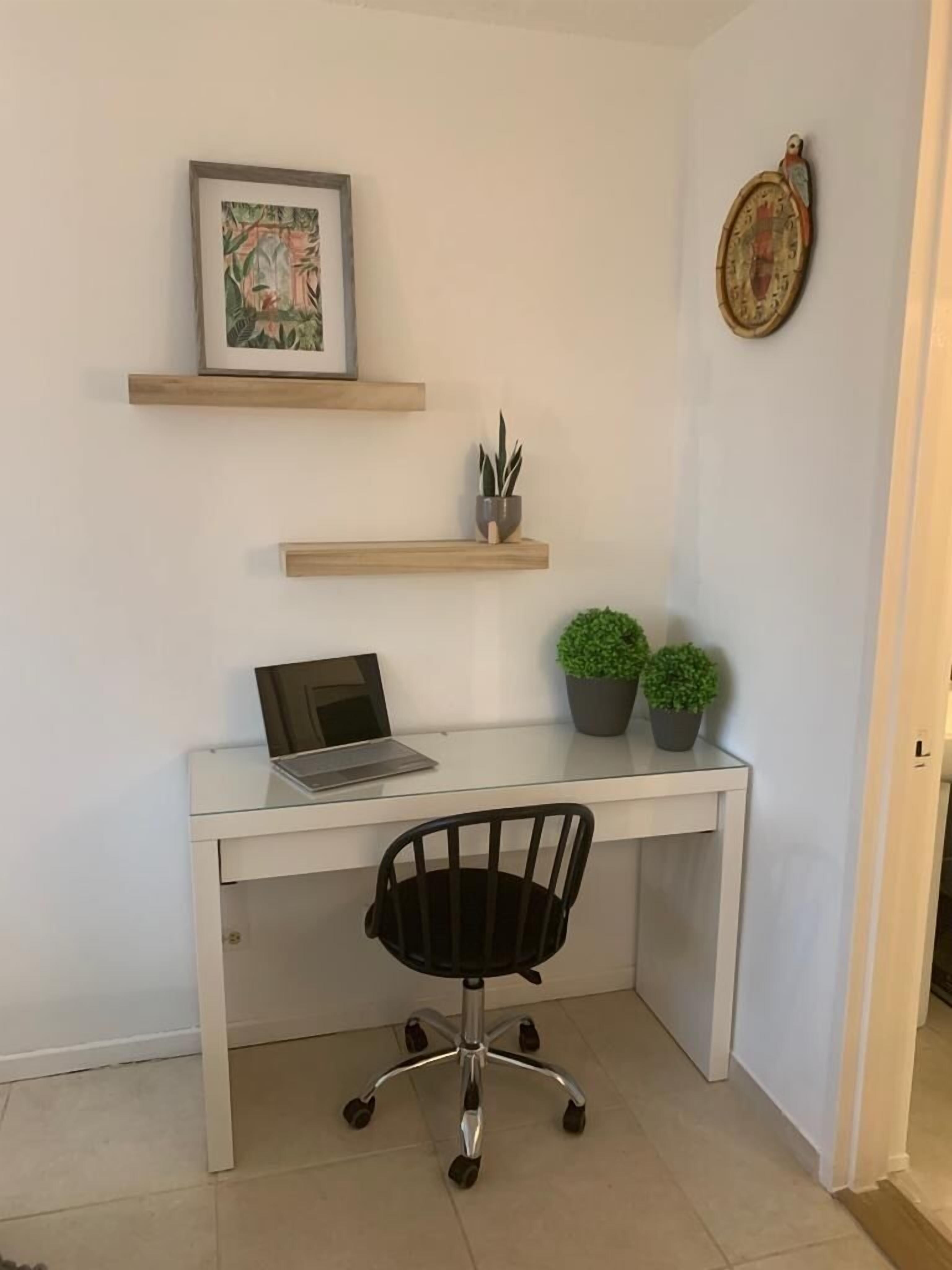 A white desk with a laptop, two potted plants, and a framed picture on a wall.
