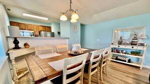 A dining area with a wooden table and chairs, a kitchen in the background, and a bookshelf with books and decorative items.