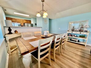 Un área de comedor con una mesa y sillas de madera, una cocina al fondo y una estantería con libros y adornos.
