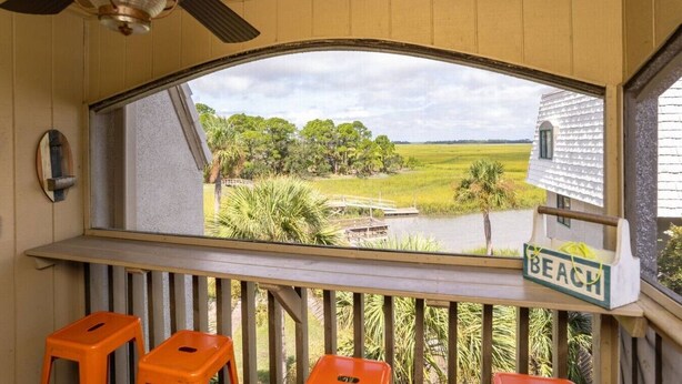 A room with a large window overlooking a beach, a ceiling fan, and orange stools.