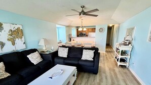 A living room with two black sofas, a white coffee table, a ceiling fan, and a kitchen in the background.