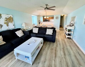 A living room with two black sofas, a white coffee table, a ceiling fan, and a kitchen in the background. - Birdie`s Beach Club Villa (resort cards available) (Saint Helena Island)