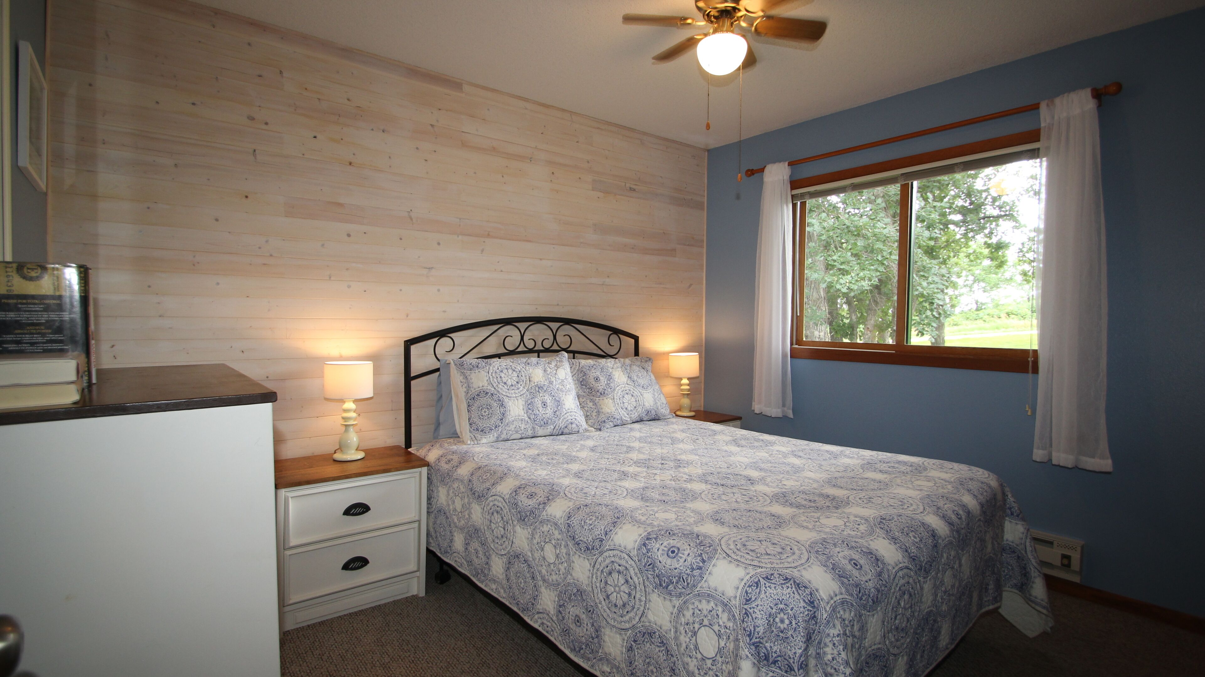 A bedroom with a bed, two bedside tables with lamps, a ceiling fan, a window with curtains, and a wooden wall.