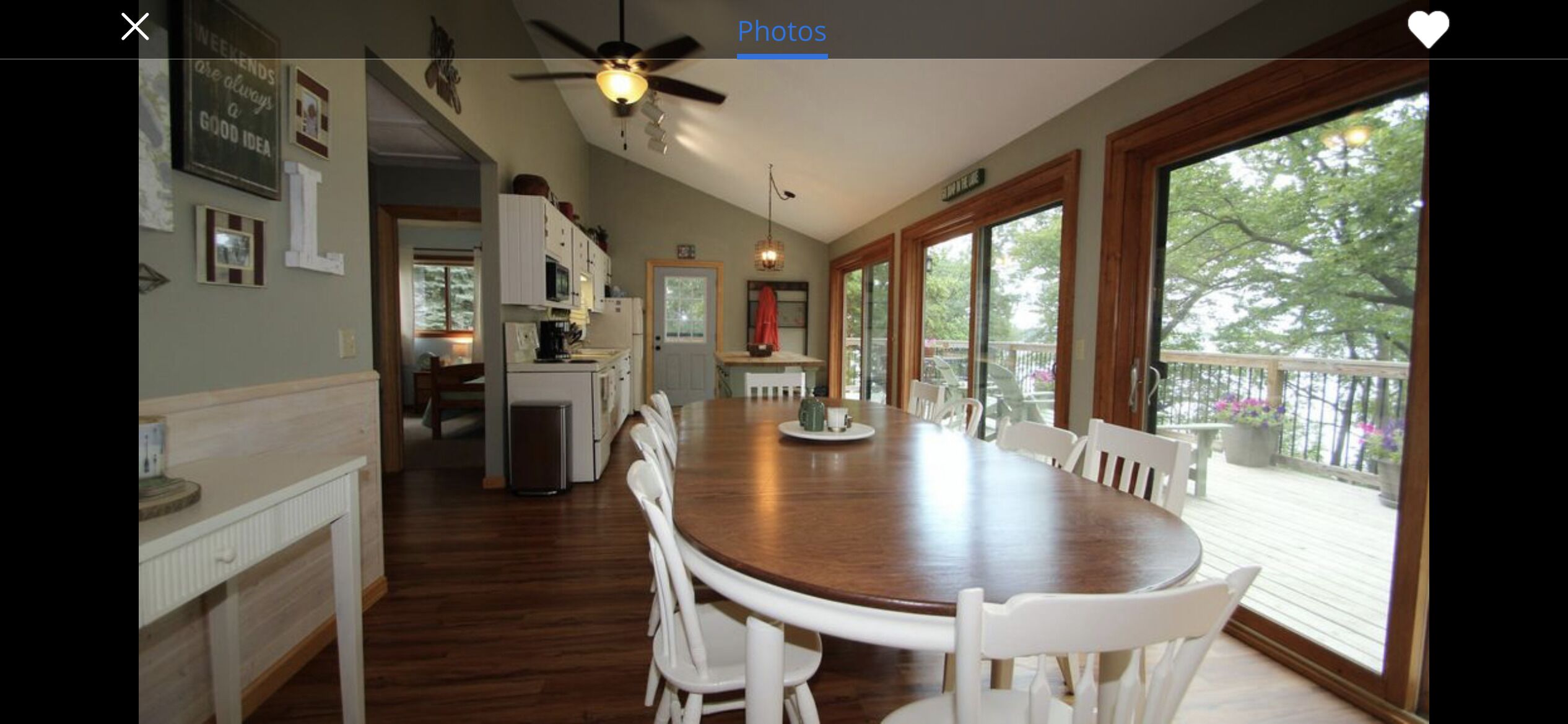 A spacious kitchen with a large wooden table and white chairs, a ceiling fan, and a view of a deck with potted plants.