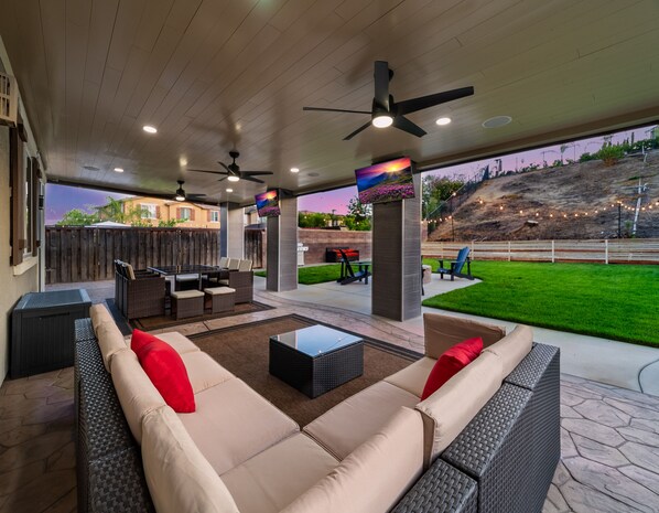 An outdoor patio area with a sofa, chairs, a table, ceiling fans, and a TV screen.