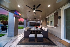 An outdoor patio with a ceiling fan, a glass-top table surrounded by wicker chairs and ottomans, and a view of a backyard.