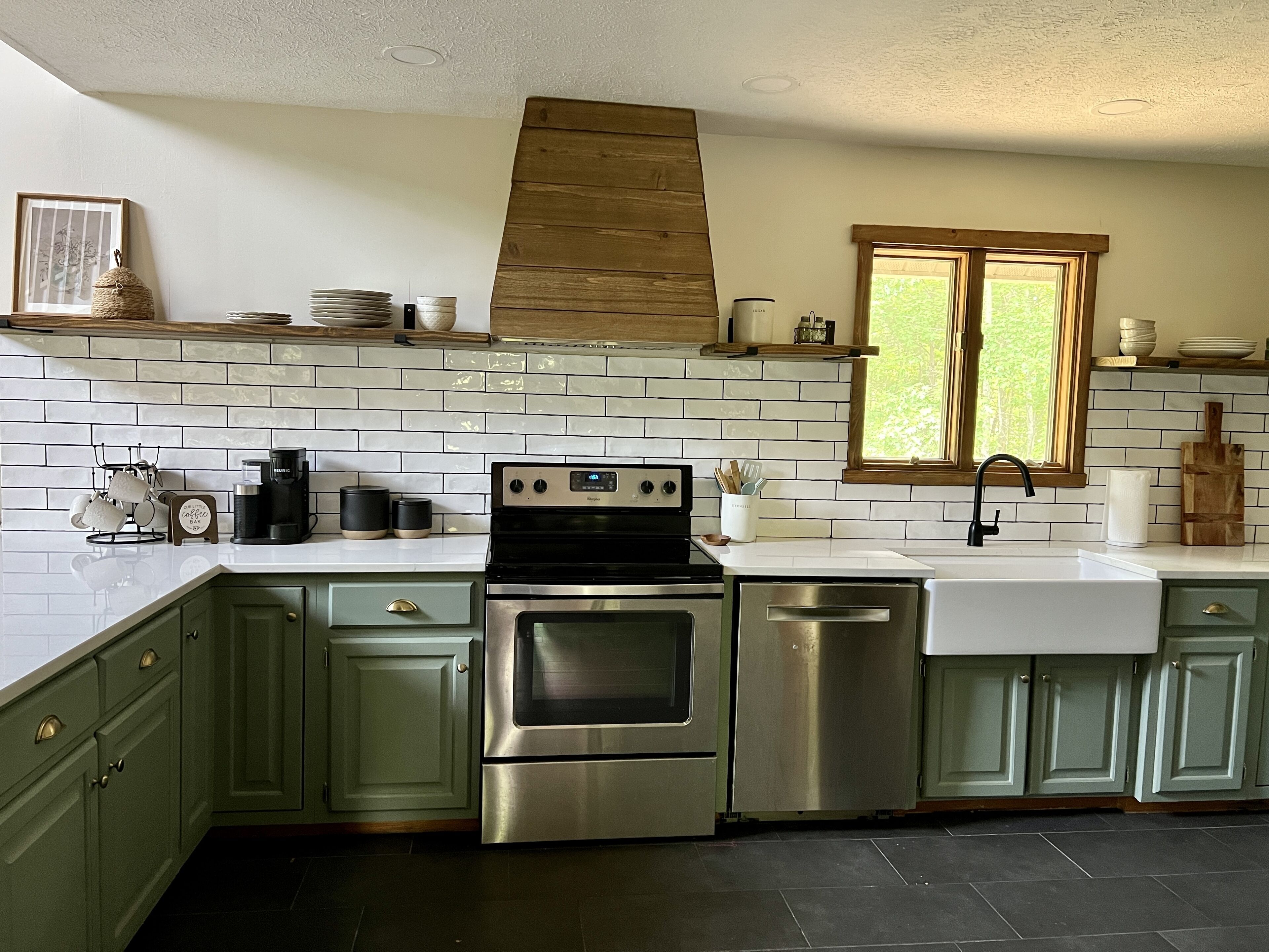 A kitchen with green cabinets, stainless steel appliances, a white tiled backsplash, and a window with a view of greenery.