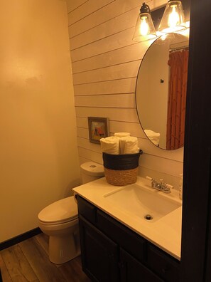 Un baño con pared de madera, un inodoro, un lavabo con espejo y una cesta de toallas.