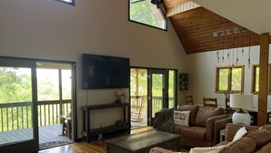 Un vaste salon doté d’un plafond en bois, d’un téléviseur à écran plat, d’une cheminée et d’une grande fenêtre donnant sur une terrasse.