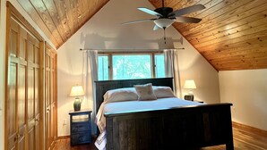 Une chambre avec un plafond en bois, un lit doté d’une tête de lit, deux tables de chevet avec des lampes et une fenêtre avec des rideaux.