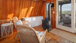 Una habitación acogedora con paredes de madera, un estufa de leña negra, sillones de mimbre y vistas a través de un ventanal.