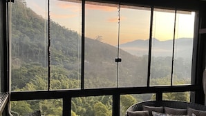 A room with a large window offering a view of a forested landscape. There are wicker chairs and a sofa with cushions.