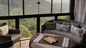 A room with a large window overlooking a mountainous landscape. There is a sofa and a chair with cushions.