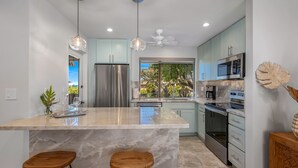 A modern kitchen with a marble island, two wooden bar stools, stainless steel appliances, and a view of the outdoors.