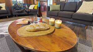 A cozy living room with a round wooden coffee table, a loaf of bread, a bottle of wine, and candles. There are two sofas with cushions and a fireplace in the background. - Mingling Waters (Nowa Nowa)