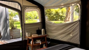 A tent with a bed, a small table with a kettle, and a window with a view of trees and a car. - Mingling Waters (Nowa Nowa)