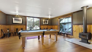 A pool table in a room with wooden walls and large windows, a fireplace, and several chairs. - Mingling Waters (Nowa Nowa)