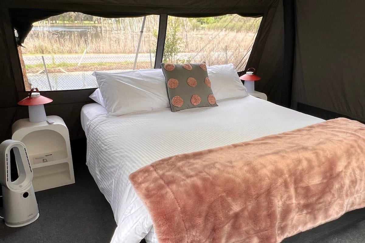 Una cama con ropa de cama blanca y una manta rosa, dos lámparas de noche y una ventana que da a una carretera y vegetación.