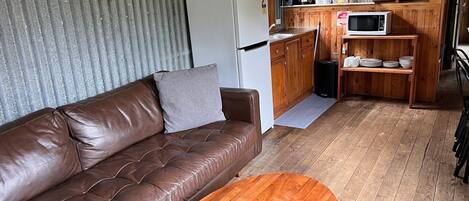 Un espacio habitable compacto con un sofá de cuero marrón, una mesa de centro de madera, una kitchenette con microondas y un refrigerador.